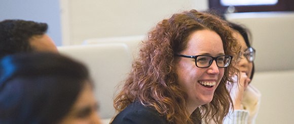 A woman with curly auburn hair laughs next to seated colleagues. 