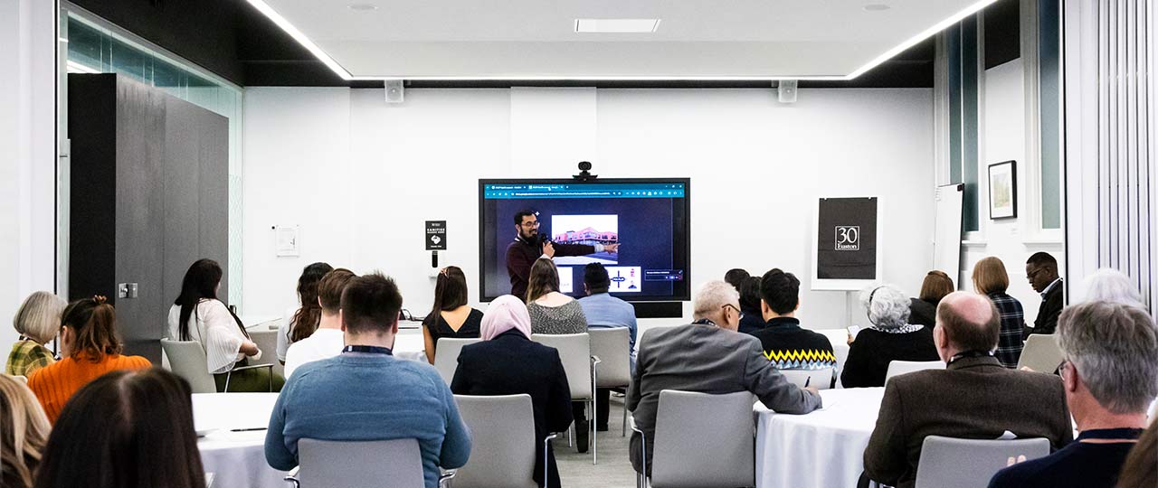 A wide, landscape image of a seated crowd looking at a presentation at the front of a large, white room. 