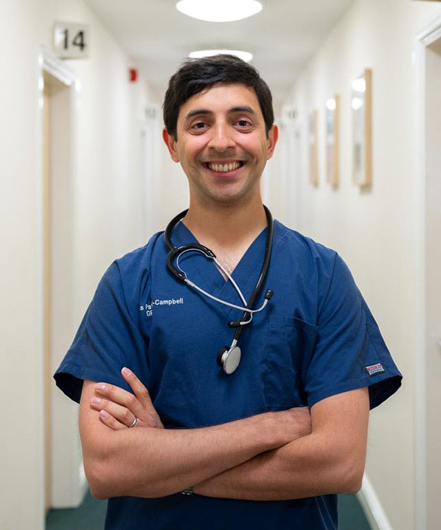 Dr Thomas Patel Campbell smiles at the camera with his arms crossed wearing blue hospital scrubs. 