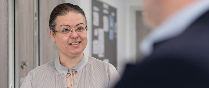 A woman with dark, greying hair, wearing glasses and a grey top smiles while in conversation with a man, who is seen from over their shoulder, out of focus.