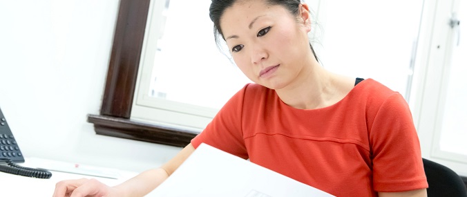 Professional woman in a red blouse sitting at a desk with paperwork in a bright office setting.