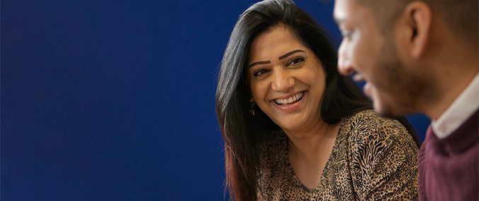 A woman with long dark hair and wearing an animal print top smiles while in conversation with a bearded man.