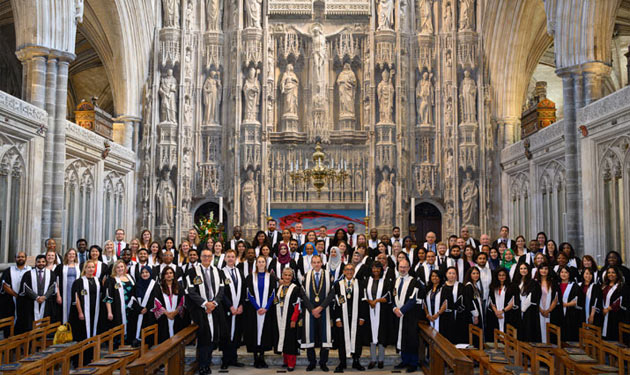 A large group of ceremony attendees wearing gowns stand together for a photo inside a large church.