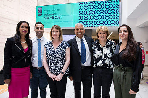 Six people stood in a line smiling. Behind them is a screen that says "welcome to the advocate summit 2025, the background is a teal green colour with rcgp advocate branding. 