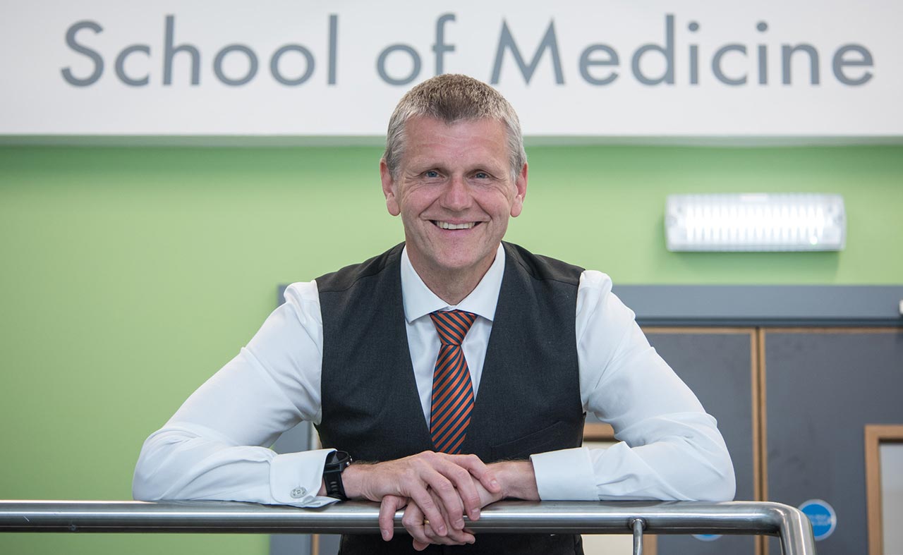 Professor Scott Wilkes leans against a railing, in front of a sign reading "School of Medicine".