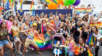 A crowd wearing rainbow clothing hold banners and balloons. 