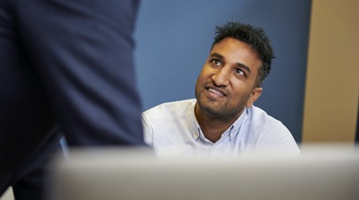 A man sitting at a desk looks up and smiles at a colleague standing up out of shot.