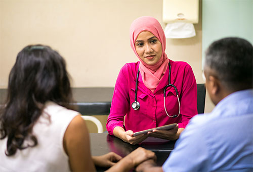 A female doctor wearing a pink hijab and stethoscope holds a tablet while speaking with two patients seated across from her in a consultation room.