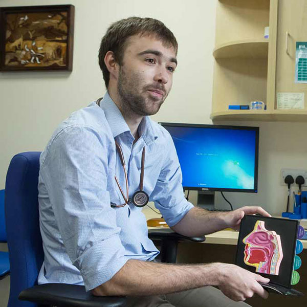 A male doctor with a stethoscope sitting at his desk, holding a tablet displaying anatomical diagrams. He's wearing a light blue shirt and is positioned in front of a computer monitor in a medical office setting.