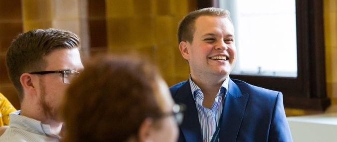 A smiling man wearing a navy suit sits with two colleagues.