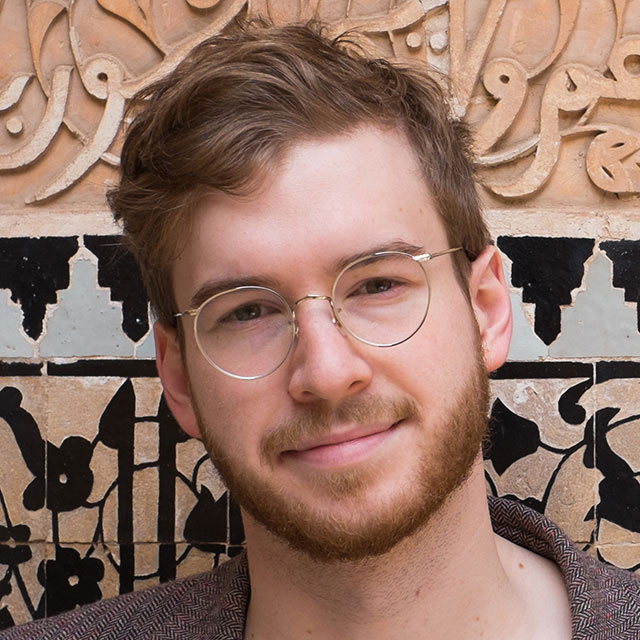 A headshot of James Morton wearing glasses in front of a decorative stone background.